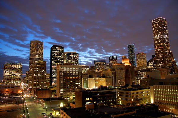 Downtown Houston skyline at dusk with glowing office buildings under a dramatic cloudy sky.